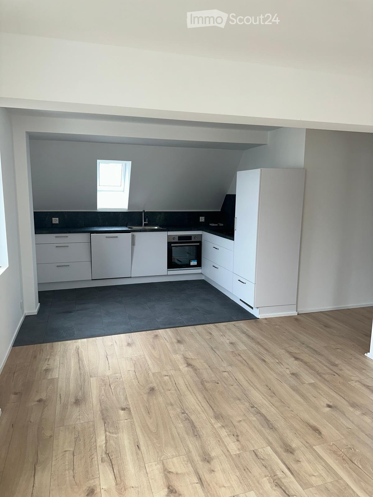Modern kitchen with white cabinets, stainless steel appliances, and a dark countertop. The floor is light wood, and there is a small window above the sink.