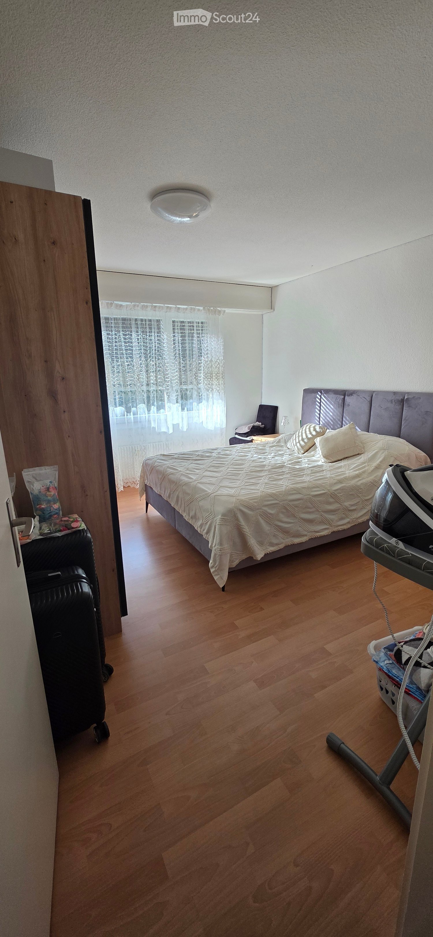A well-lit bedroom with wooden flooring, a large bed with a patterned duvet and pillows, a nightstand with a lamp, a chair, and a suitcase near the door. An ironing board and cover are placed near the bed.