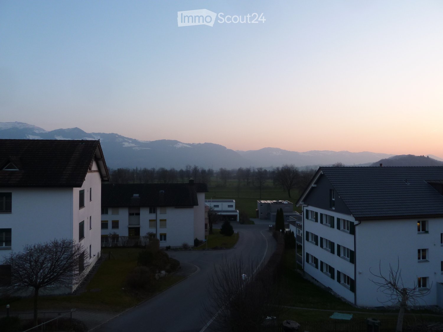 Residential complex with multiple buildings, surrounded by greenery and mountains in the background.
