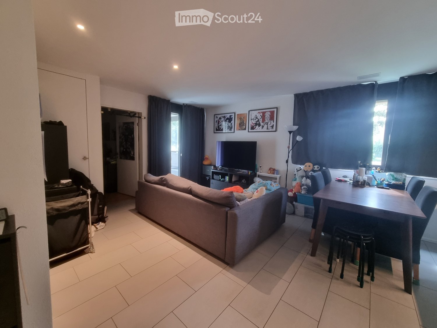 A modern living room with a beige sofa, a wooden dining table with chairs, a television, and large windows with dark curtains. The room features tiled flooring and several decorative items, including picture frames on the wall.