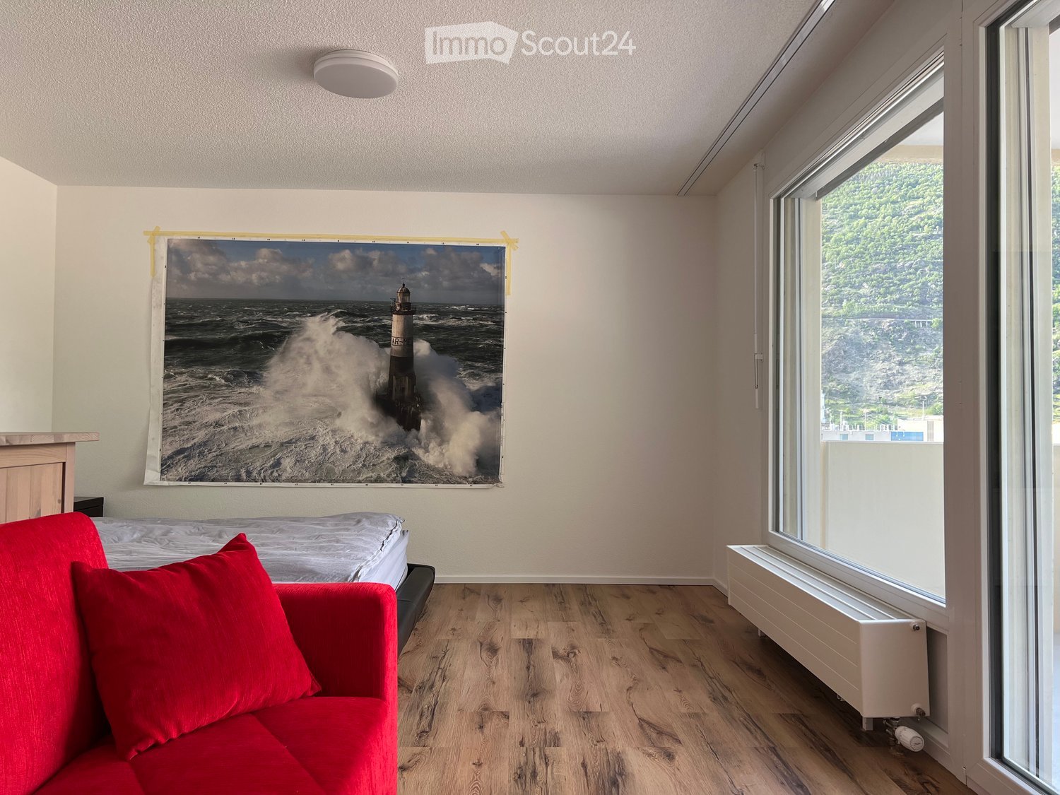 A modern, minimalist room with a red sofa, a bed, and a large picture of a lighthouse on the wall. The room features wooden flooring and large windows with a scenic view.