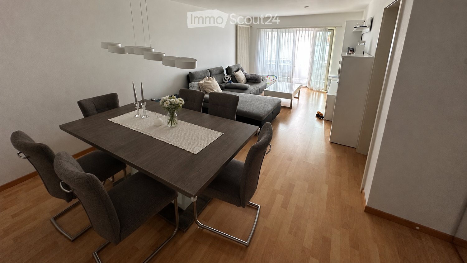 Modern living and dining area with a grey sofa, ottoman, coffee table, dining table with chairs, and a flower vase on the table.