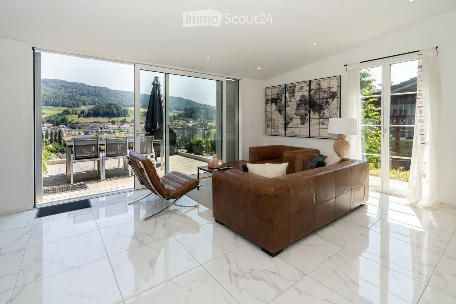 Modern living room with white marble flooring, large windows offering scenic views, leather sofa, armchair, coffee table, and decorative art piece on the wall.