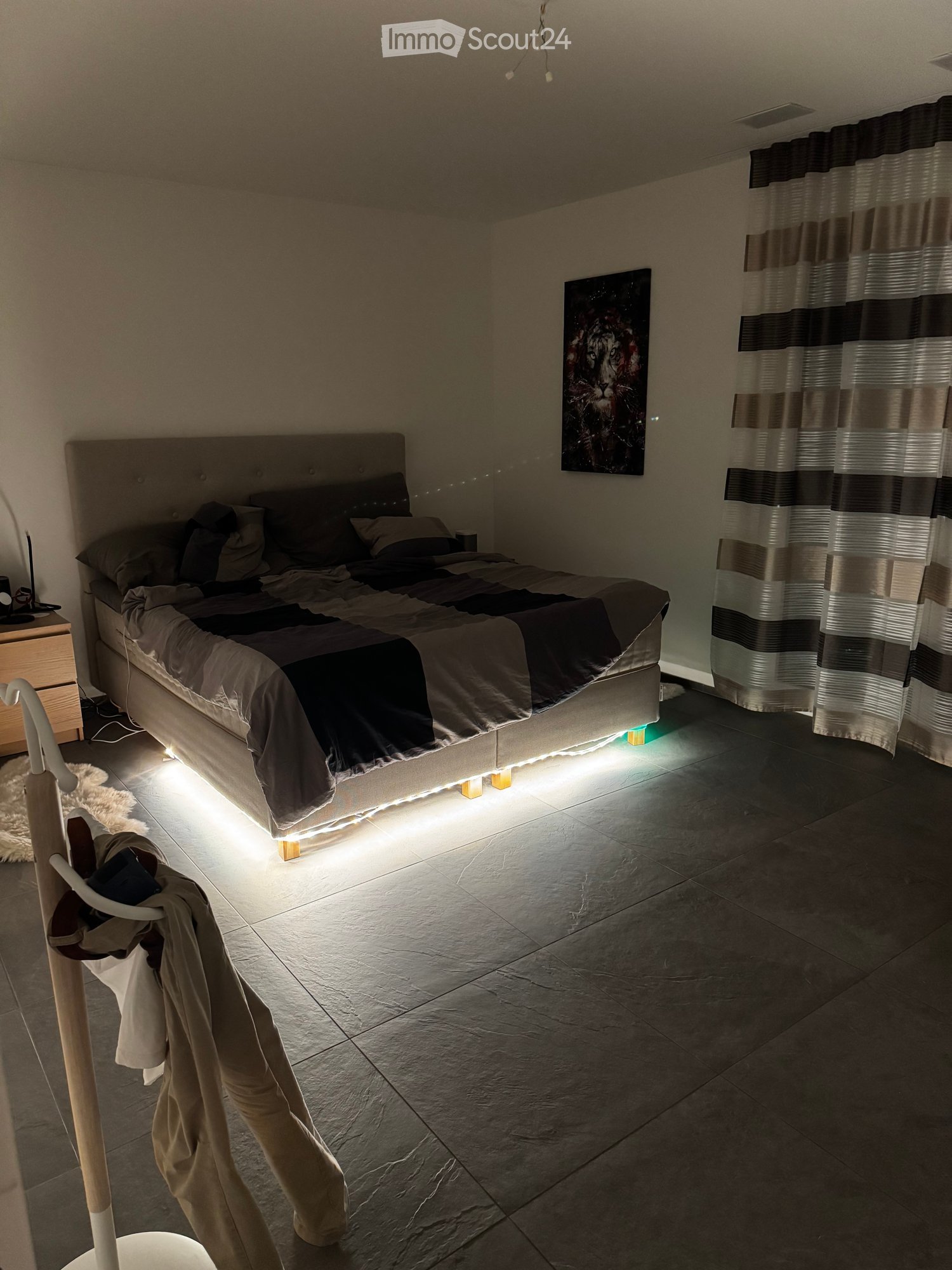 Modern bedroom with a large bed, nightstand, painting, and a curtained window. The room features tiled flooring and ambient lighting under the bed.