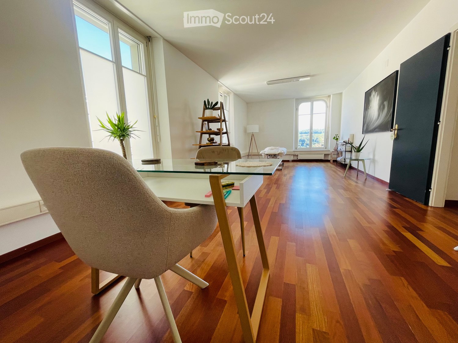 Bright, modern room with wooden flooring, large windows, a glass table, a comfortable chair, a shelf with plants, and a screen on the wall.