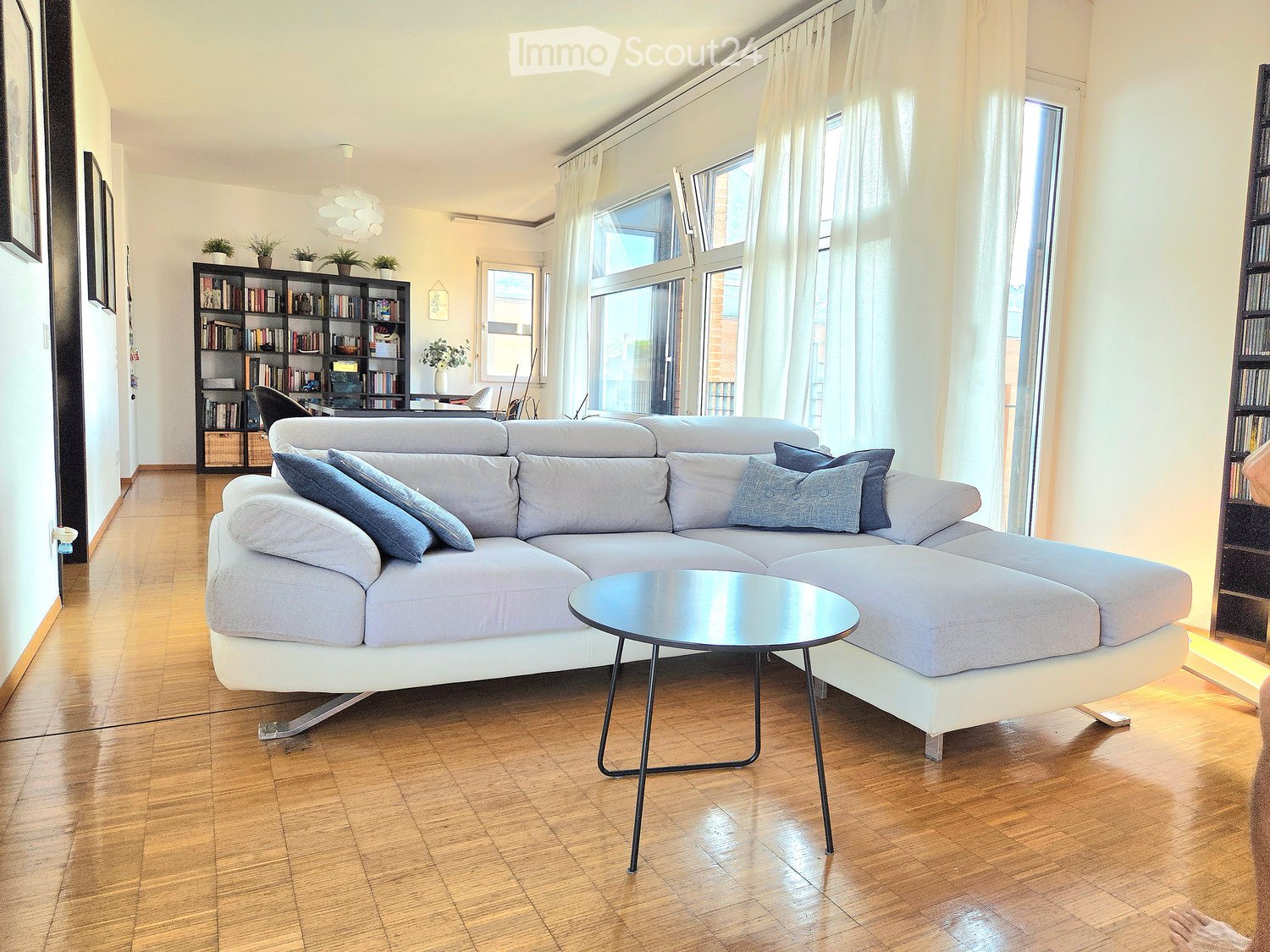 Modern living room with a grey sectional sofa, coffee table, bookshelf, and large windows with white curtains.