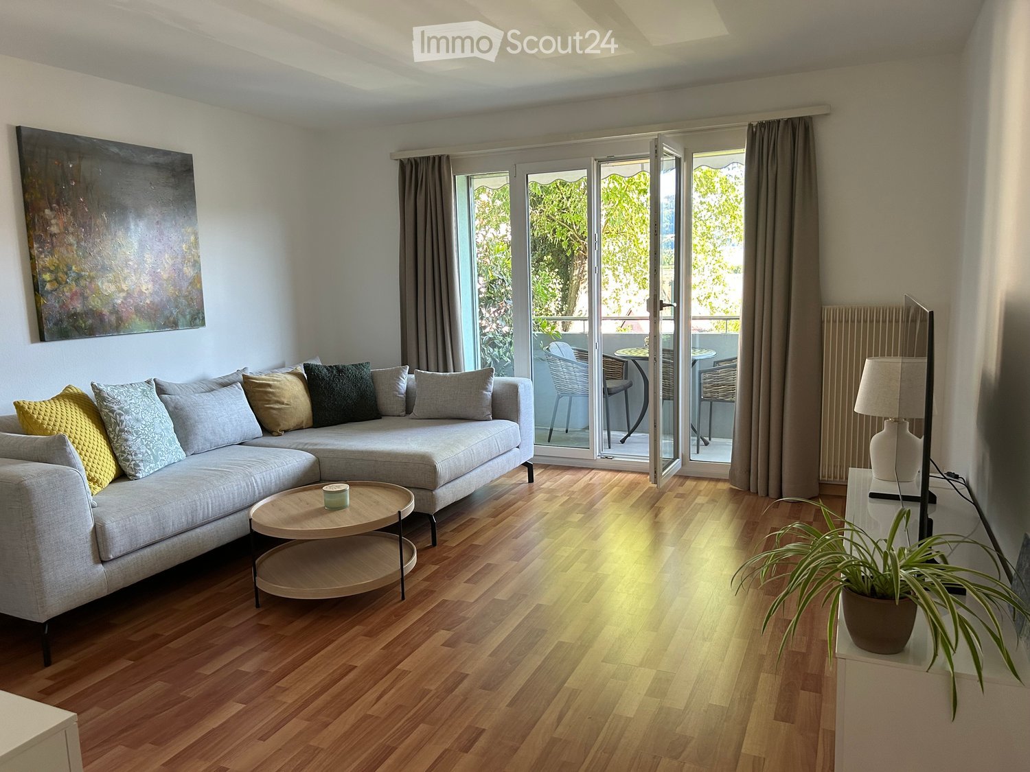 Modern living room with a grey sectional sofa, wooden coffee table, table lamp, potted plant, and a view of a balcony with outdoor furniture.