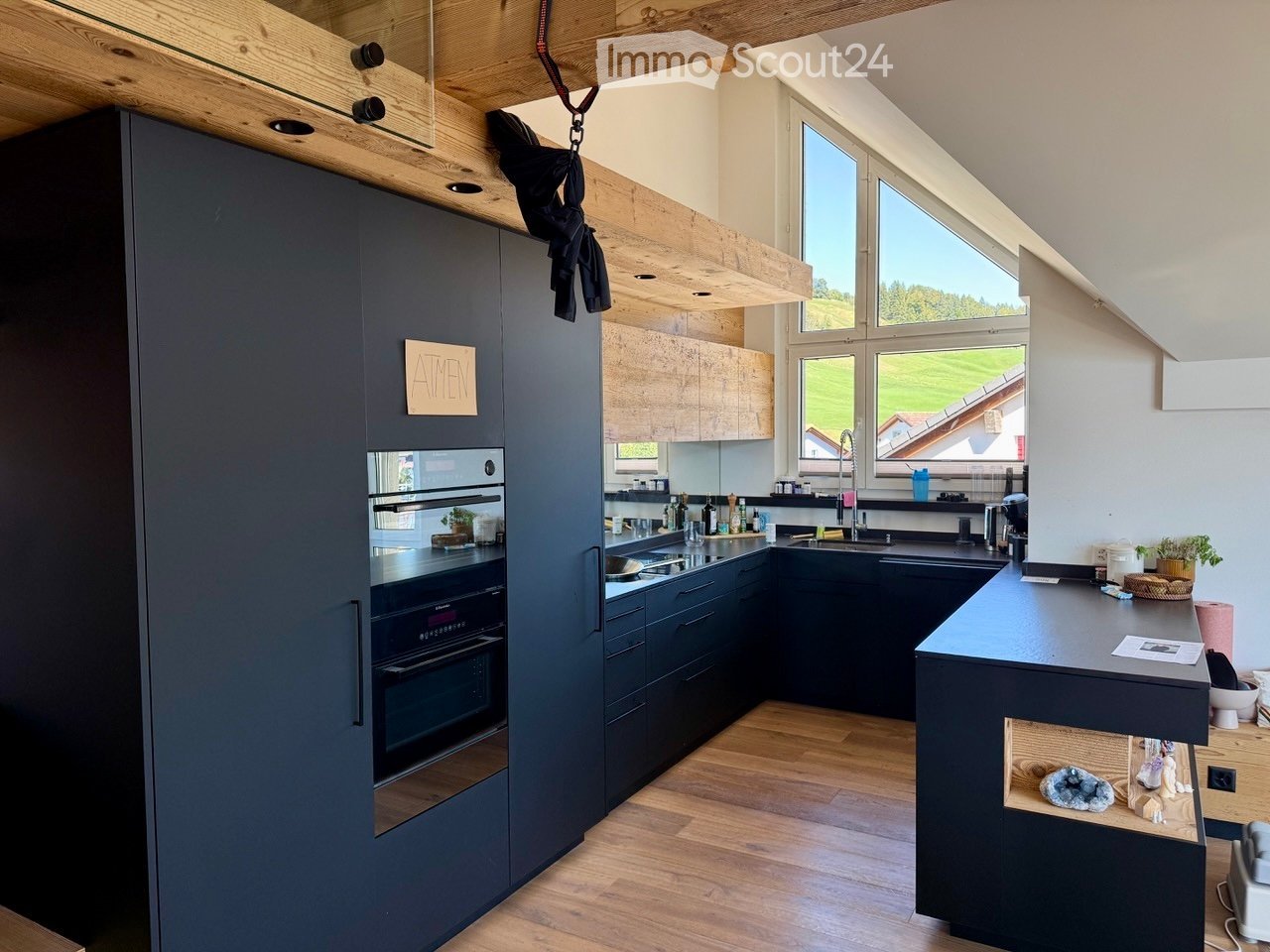 Modern kitchen with dark grey cabinets, wooden flooring, and large windows overlooking a green landscape. Features include an oven, sink, and countertop.