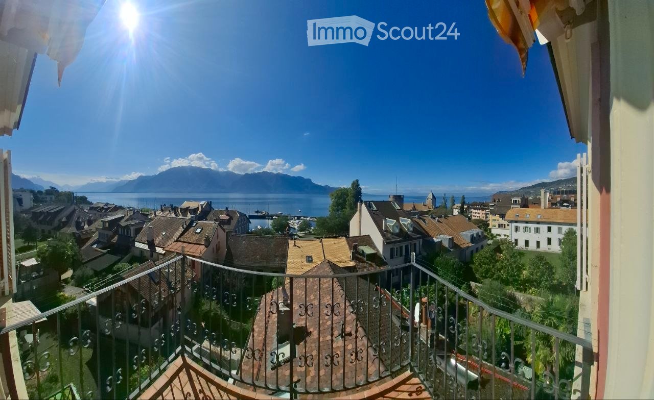 Balcony overlooking a town with a lake and mountains in the background, featuring a metal railing and wooden decking.