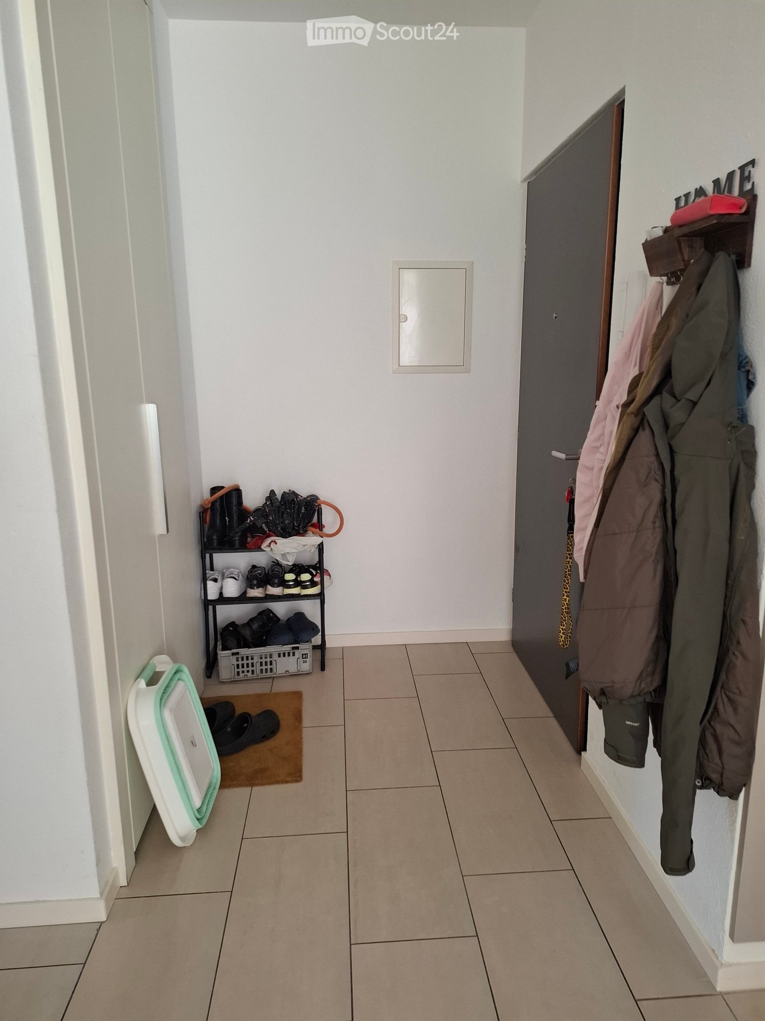A narrow hallway with tiled flooring, white walls, and a coat rack holding jackets and shoes. A shoe shelf with multiple pairs of shoes is visible on the left side.