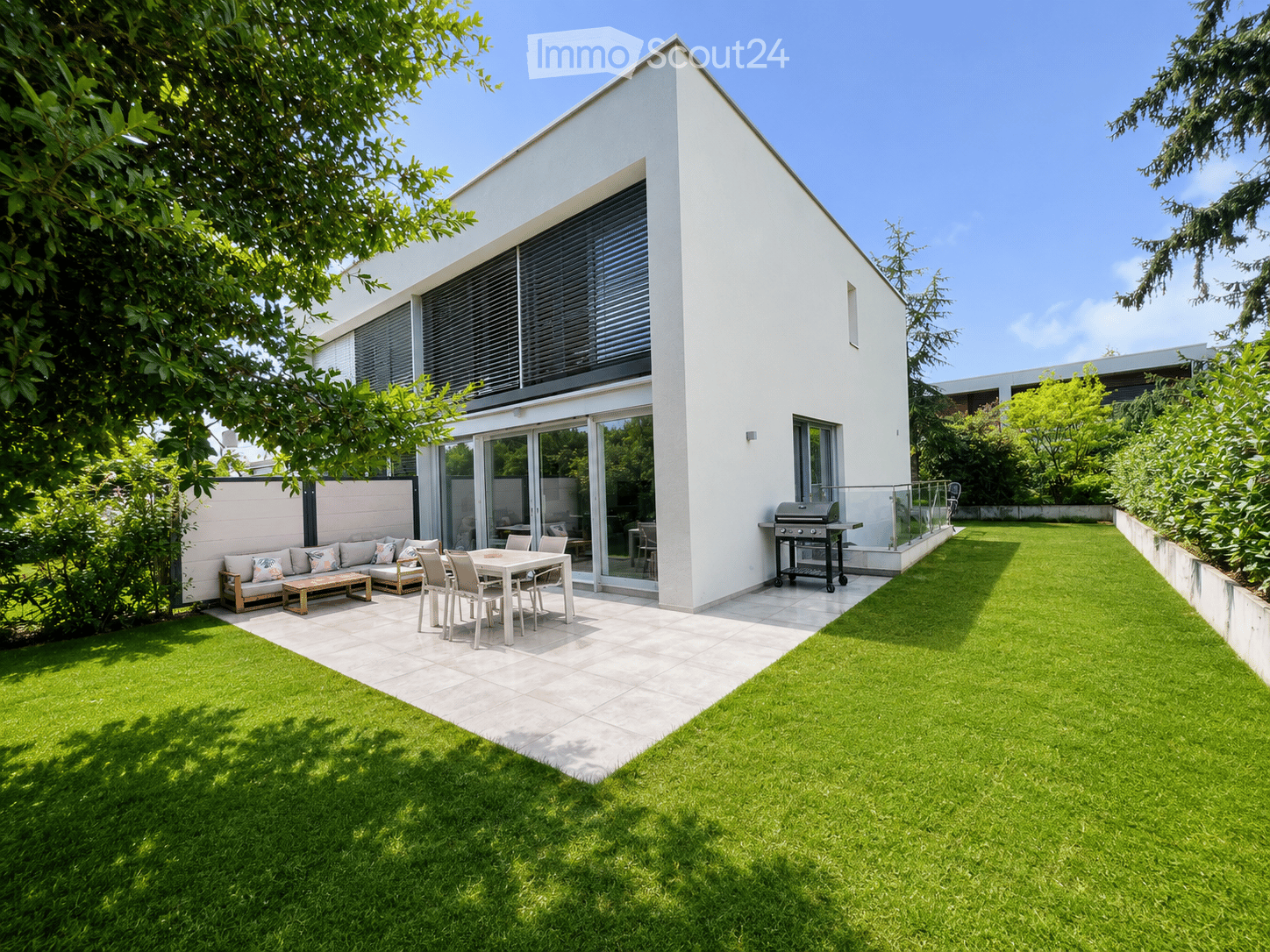 Modern outdoor space with a well-maintained lawn, patio area, and a contemporary house with large windows and a sliding glass door. Features include a sofa, coffee table, dining table with chairs, and a grill.