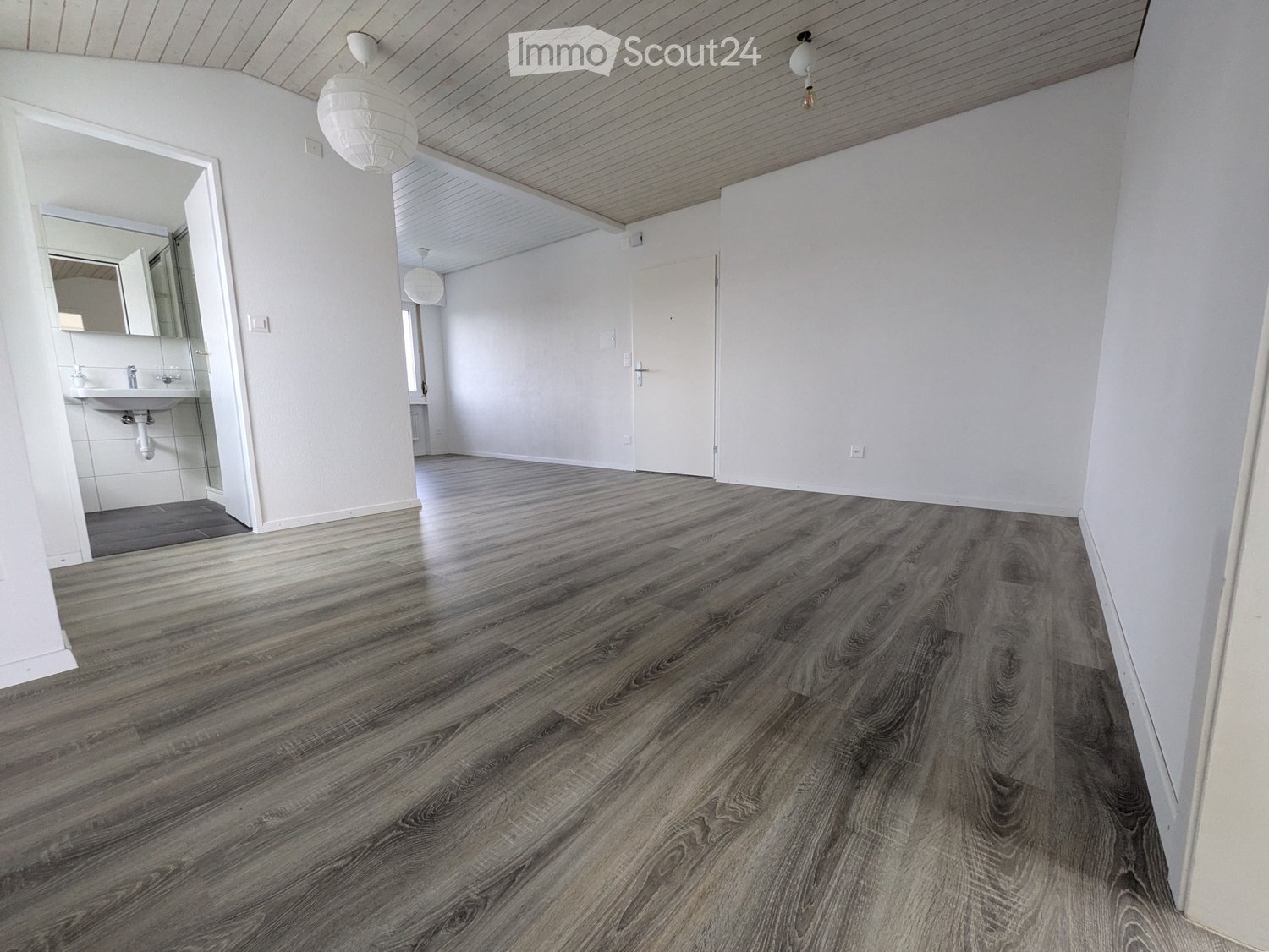 Empty room with wooden flooring, white walls, and a partially visible bathroom area with a sink and mirror.