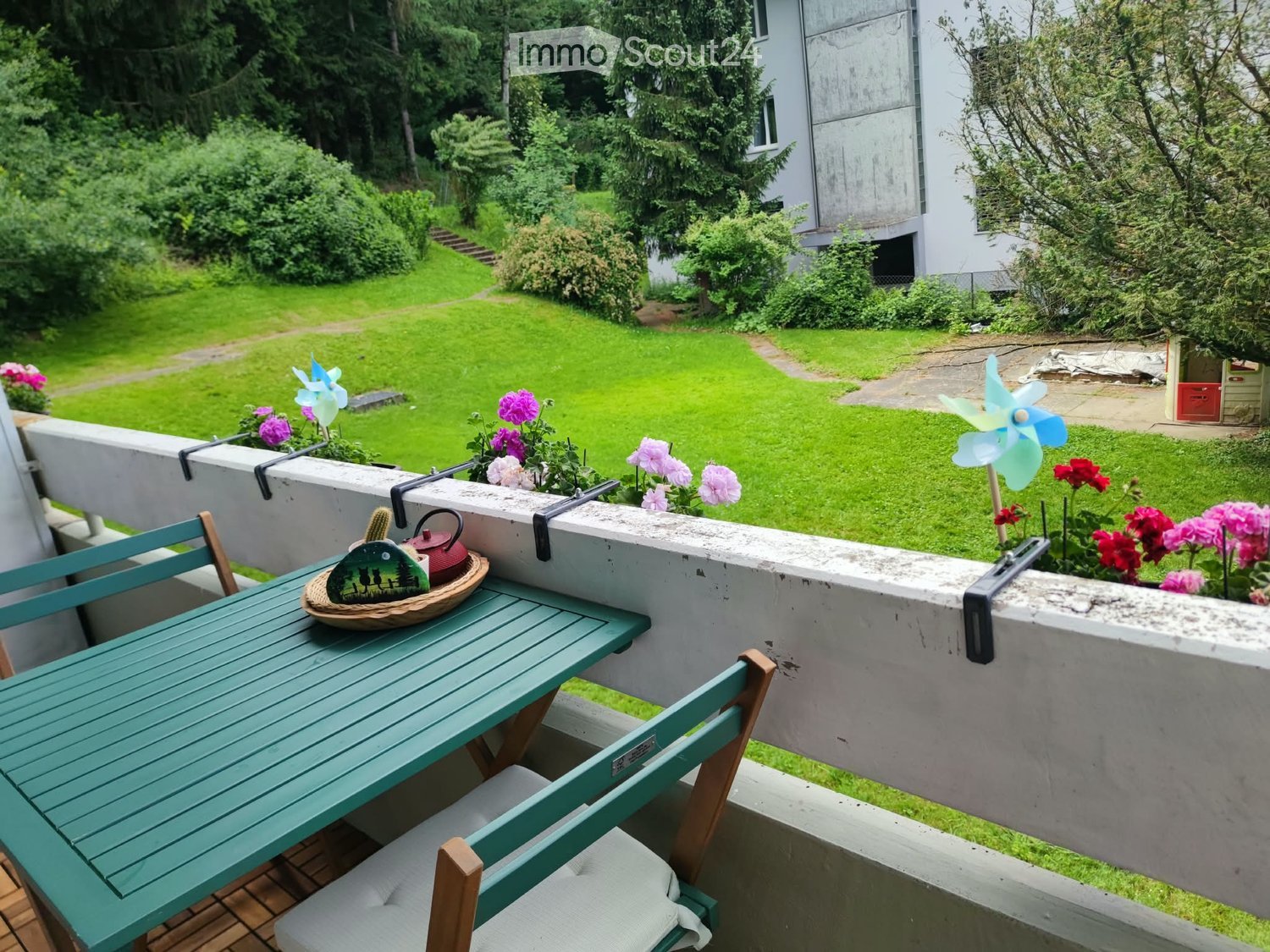 Balcony with a green table, two chairs, a teapot, and a wicker basket, overlooking a well-maintained garden with flowers and a windmill toy.