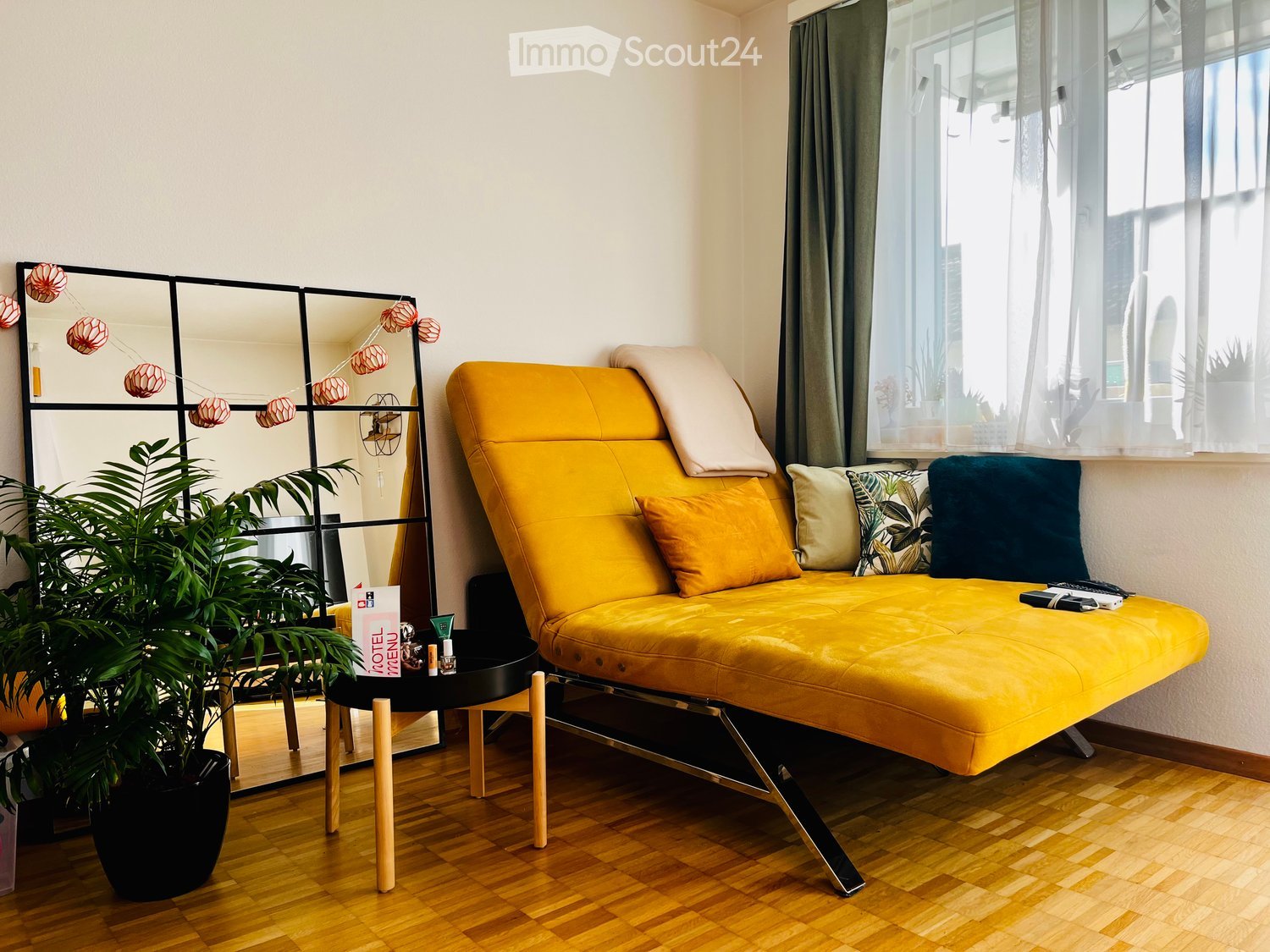 Bright, modern room with a yellow sofa, wooden floor, large window with sheer curtains, and a black side table with a potted plant.