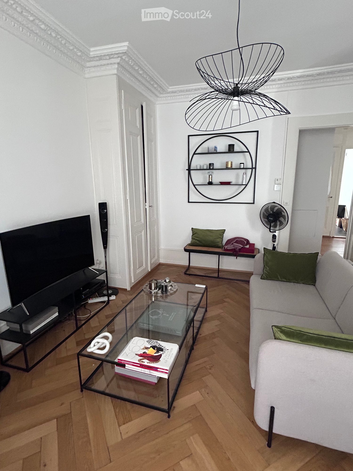 Modern living room with light-colored walls, wooden flooring, and a grey sofa with green cushions. A glass coffee table holds books and decorative items, and a wall-mounted shelf displays small objects. A television and speaker system are on the left side