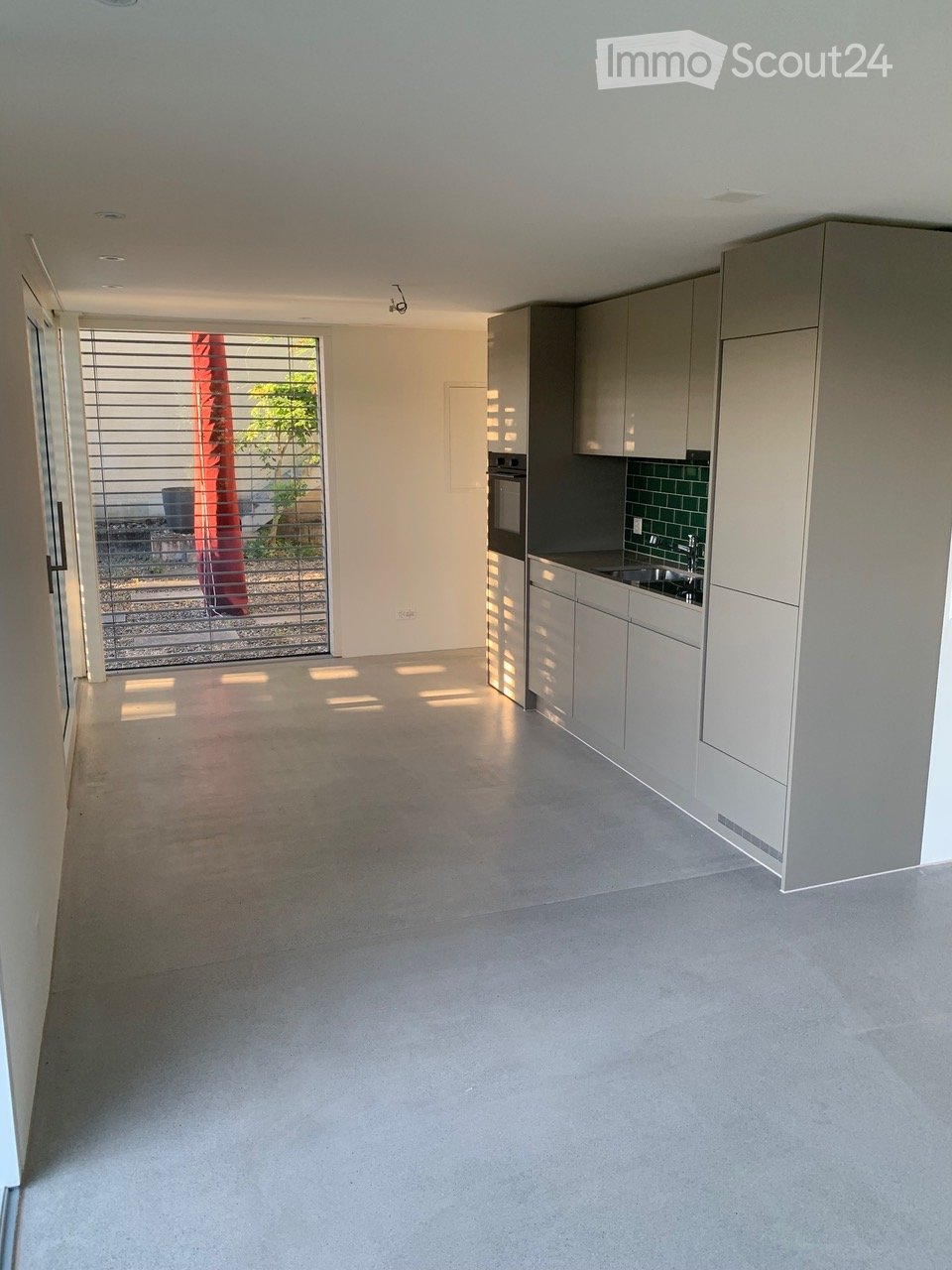 Empty kitchen with modern grey cabinets, stainless steel appliances, and a large window with blinds leading to an outdoor area.