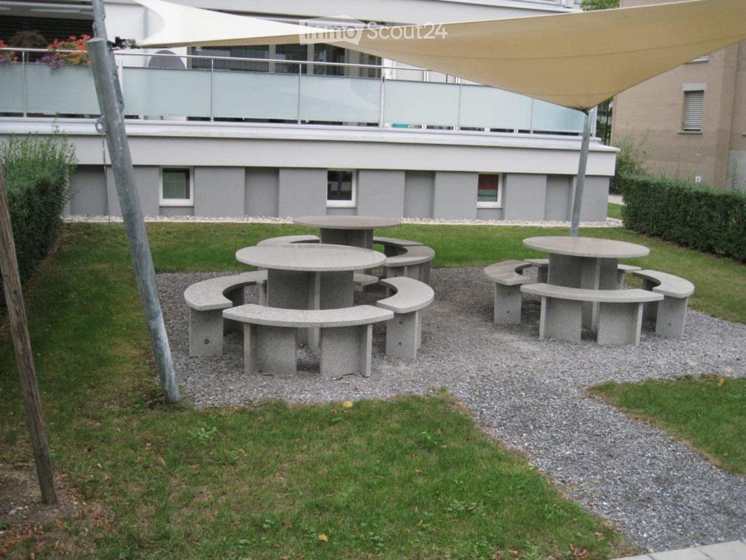 Outdoor seating area with concrete tables and benches on gravel, surrounded by grass and a modern building with a balcony.