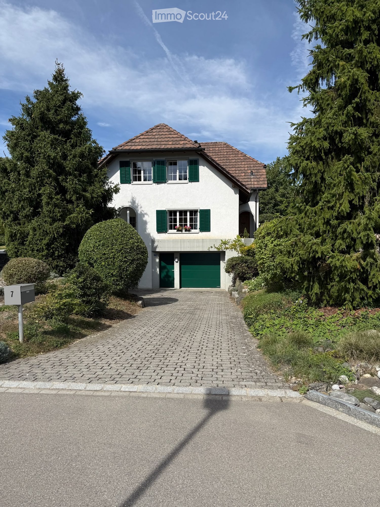 Two-story house with white exterior, green shutters, and a green garage door. The house has a tiled roof and is surrounded by trees and shrubs.