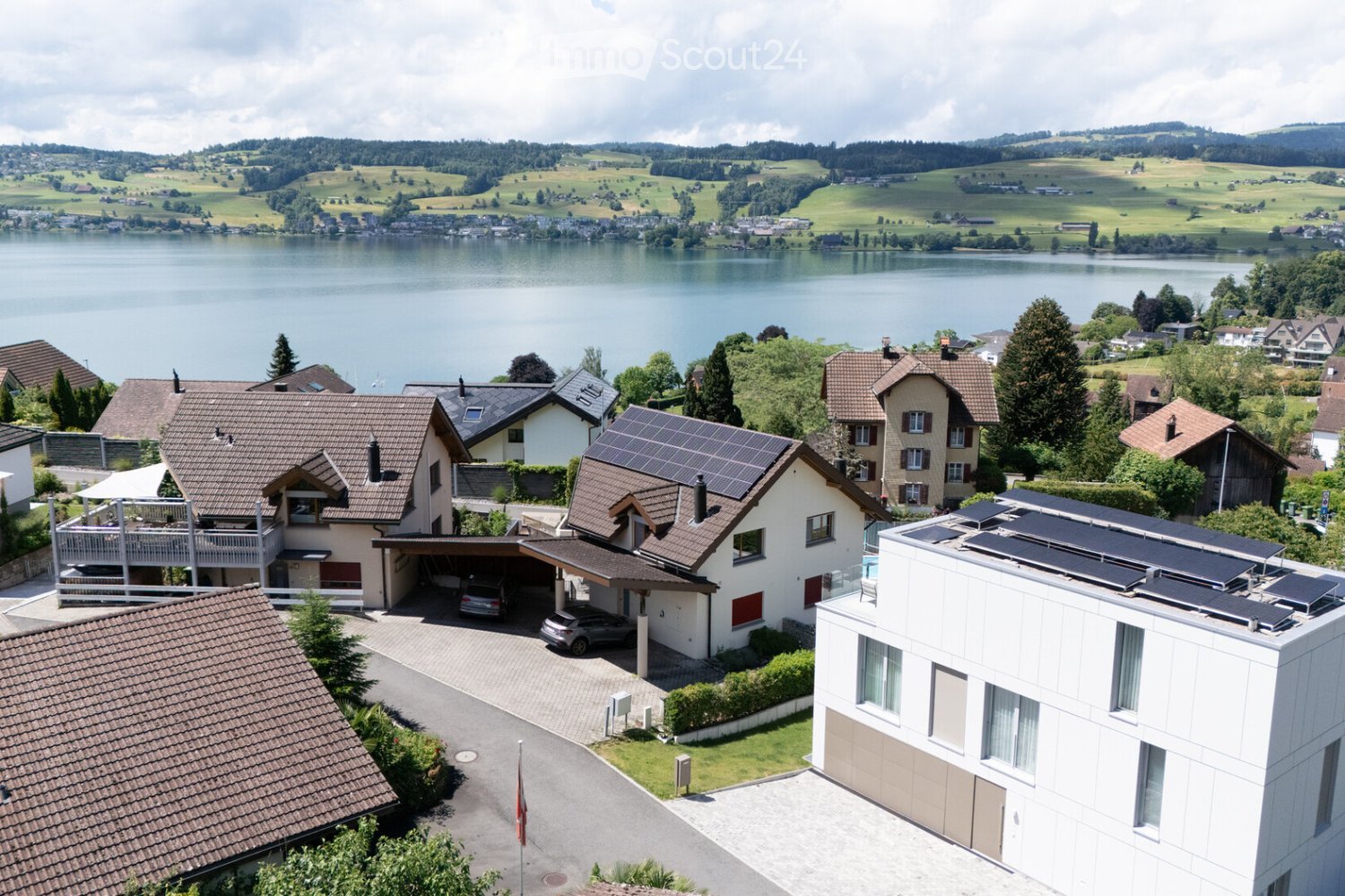 Aerial view of a residential area with multiple houses, solar panels, a lake, and surrounding greenery.