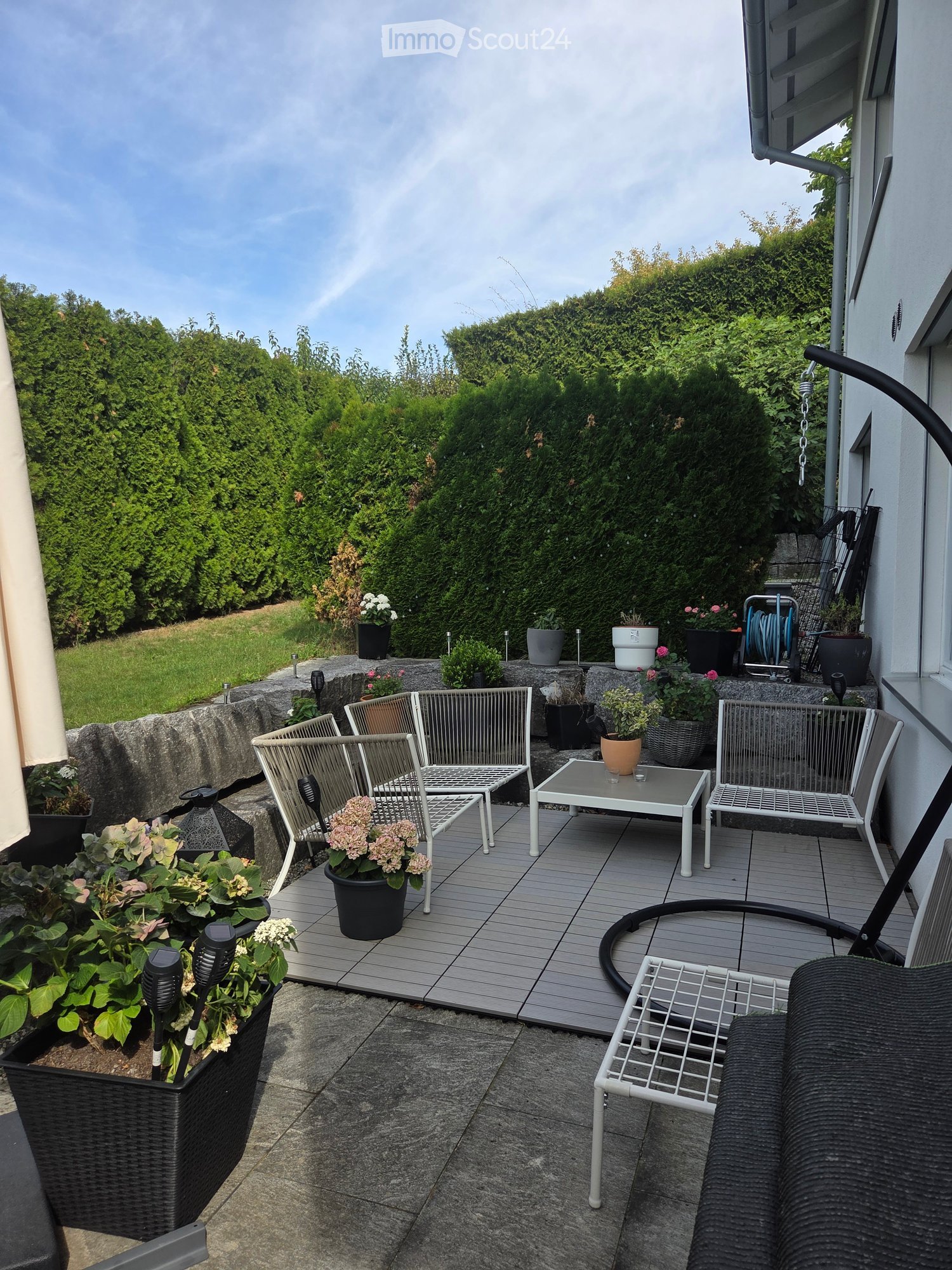 Balcony with wooden decking, white metal chairs, a small table, and potted plants.