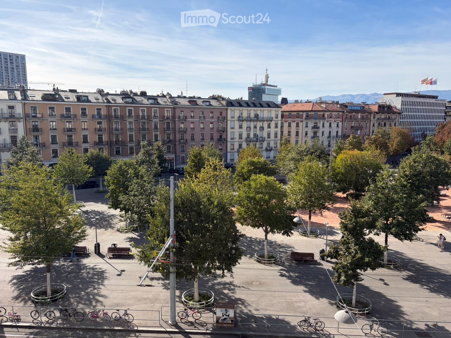 A spacious urban square with trees, benches, and bicycles parked along the edges, surrounded by multi-story residential buildings.