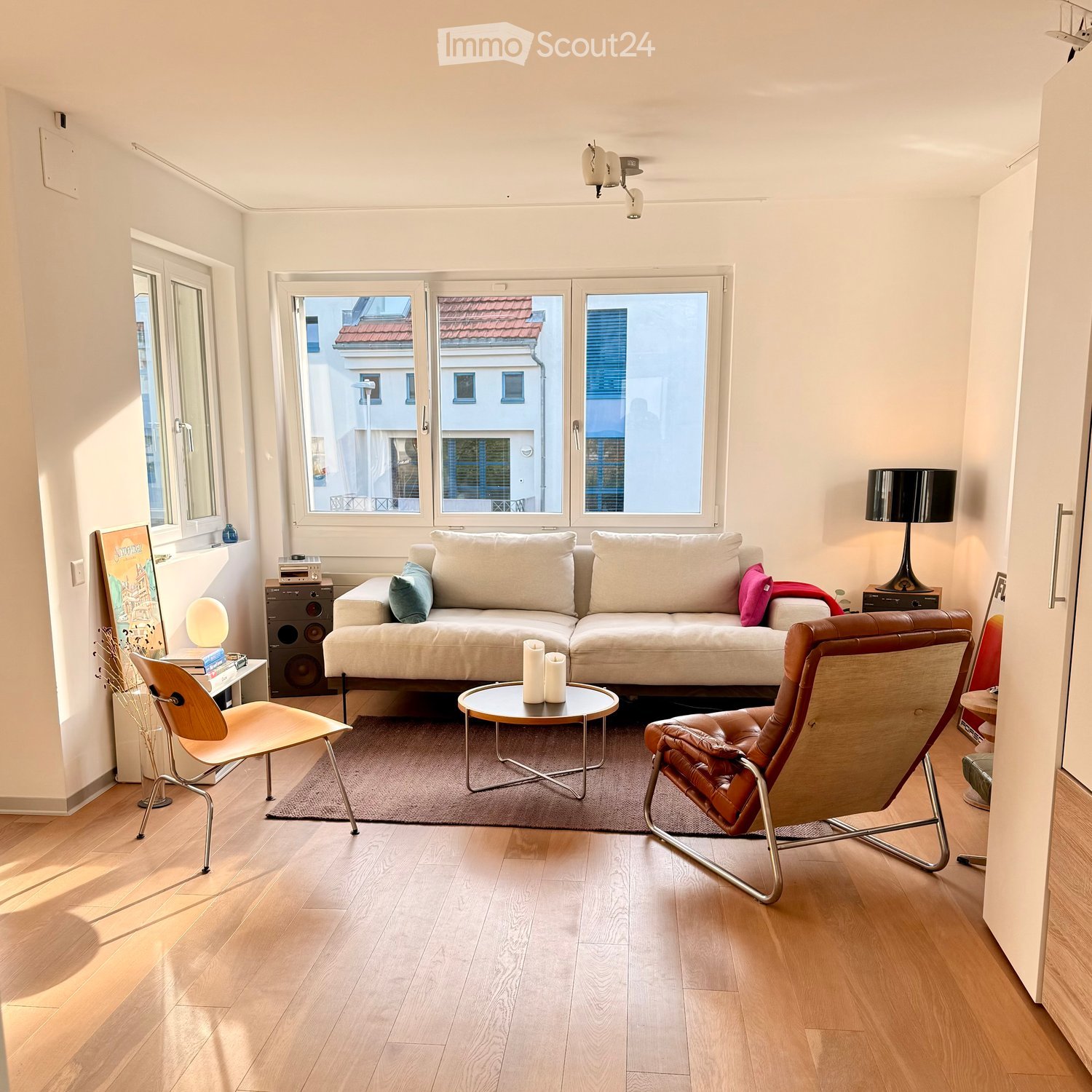Modern living room with light-colored sofa, coffee table, two chairs, and large windows offering a view of the city.