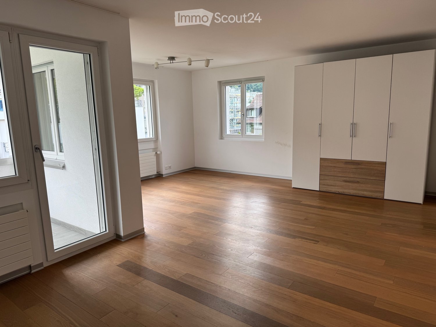 Empty room with wooden flooring, white walls, large windows, and built-in wardrobes.