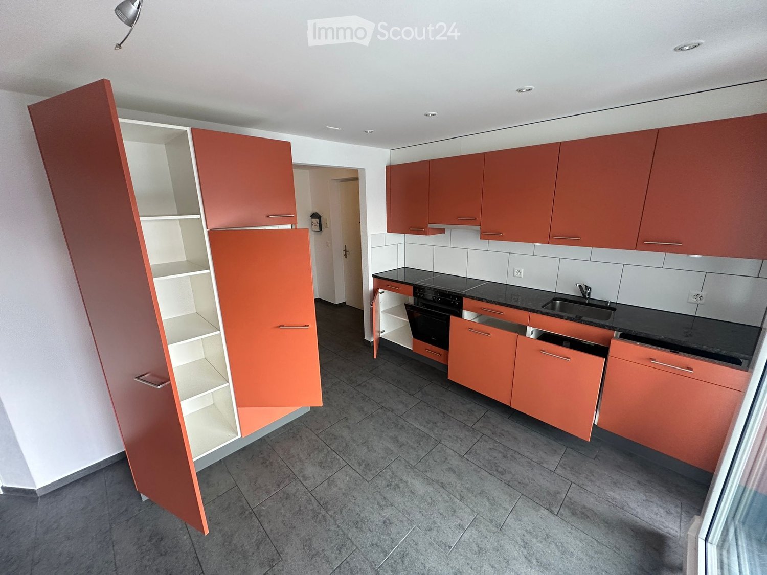 Modern kitchen with red cabinets, black countertops, and stainless steel appliances. Open shelving unit on the left side.