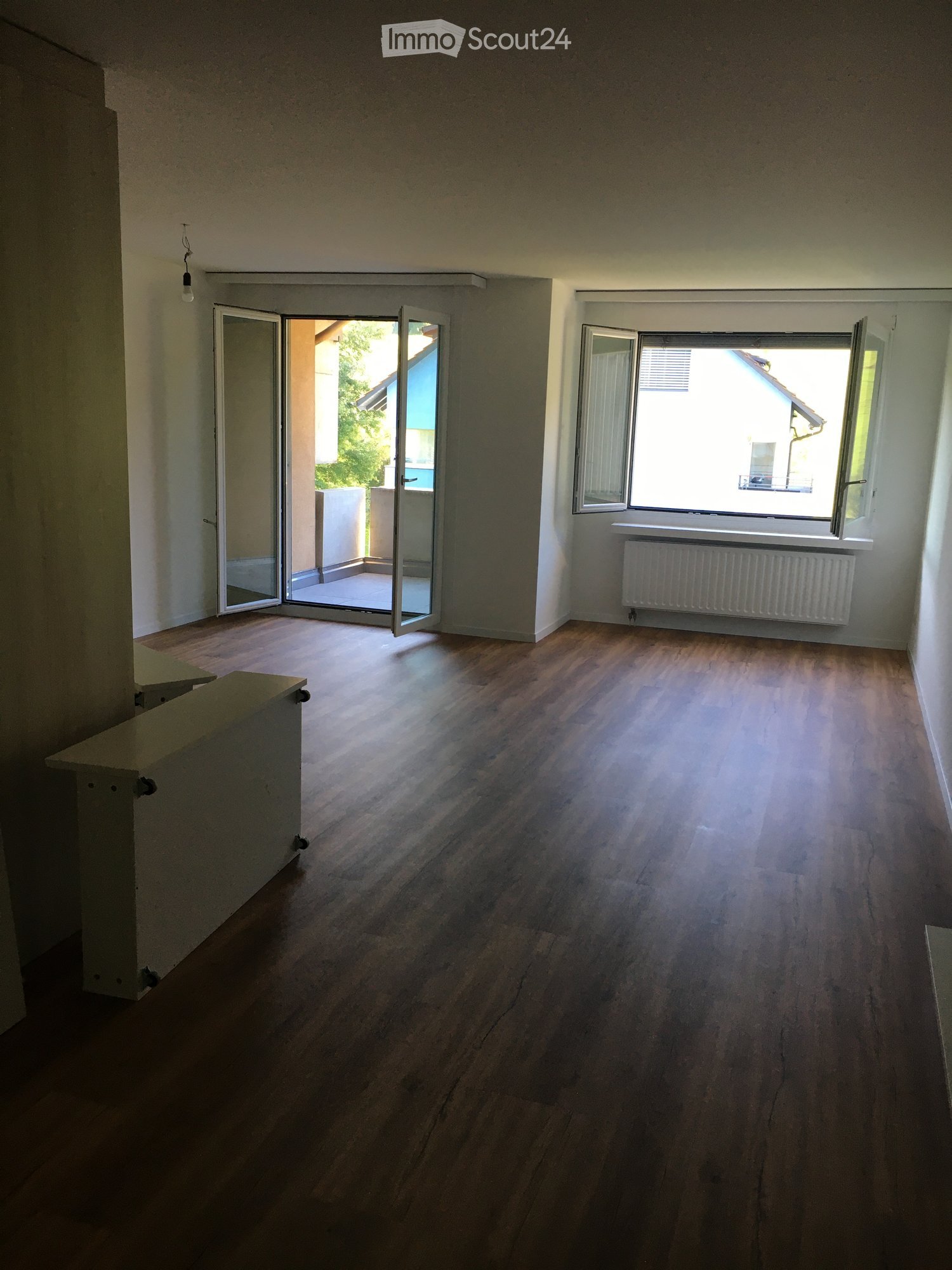 Empty room with wooden flooring, large windows, and an open sliding door leading to a balcony area.