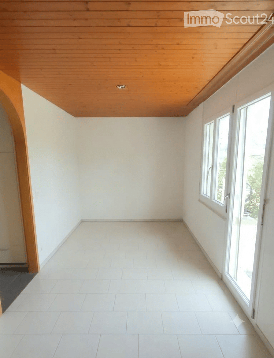 Empty room with white tiled floor, white walls, wooden slanted ceiling, and large windows on one side.