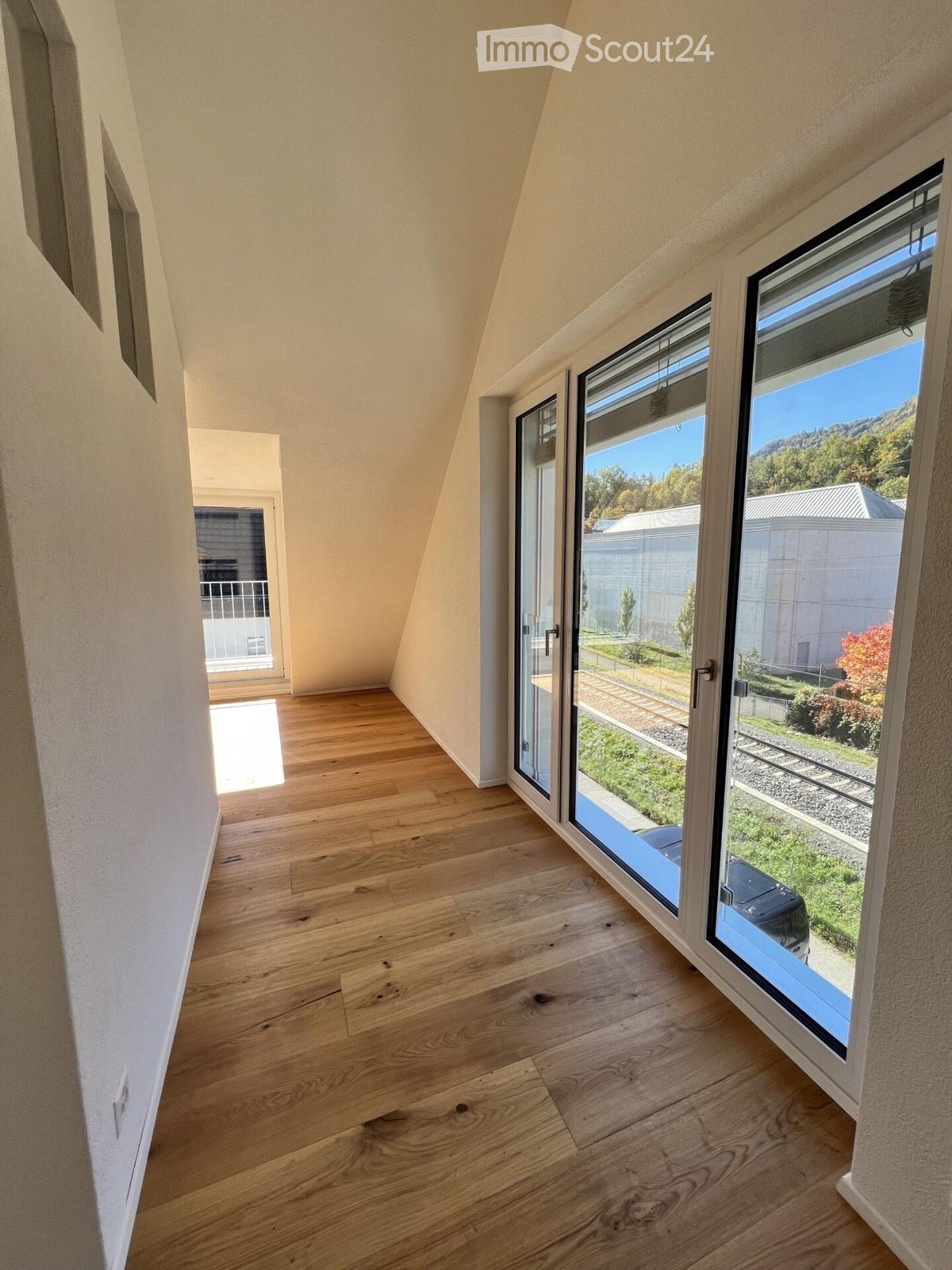 An empty room with wooden flooring and large windows offering a view of the outdoors, including a grassy area and a parked car.