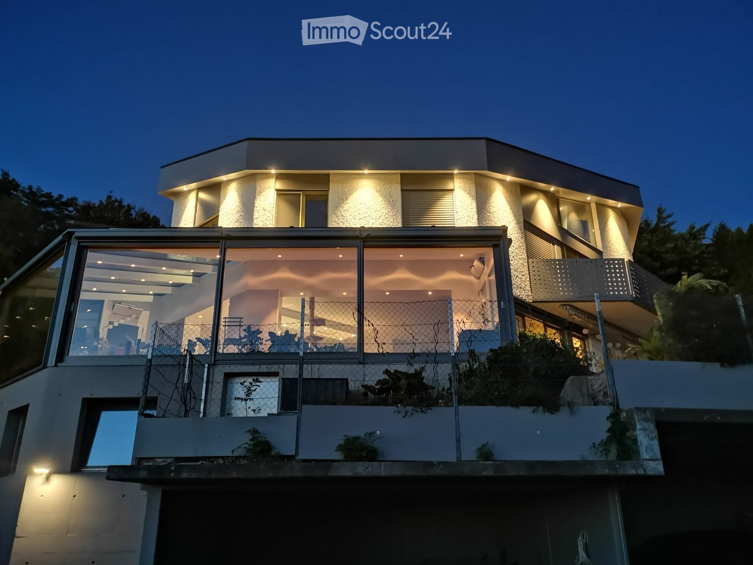 Modern two-story house with illuminated facade, large glass windows, and a raised ground floor.