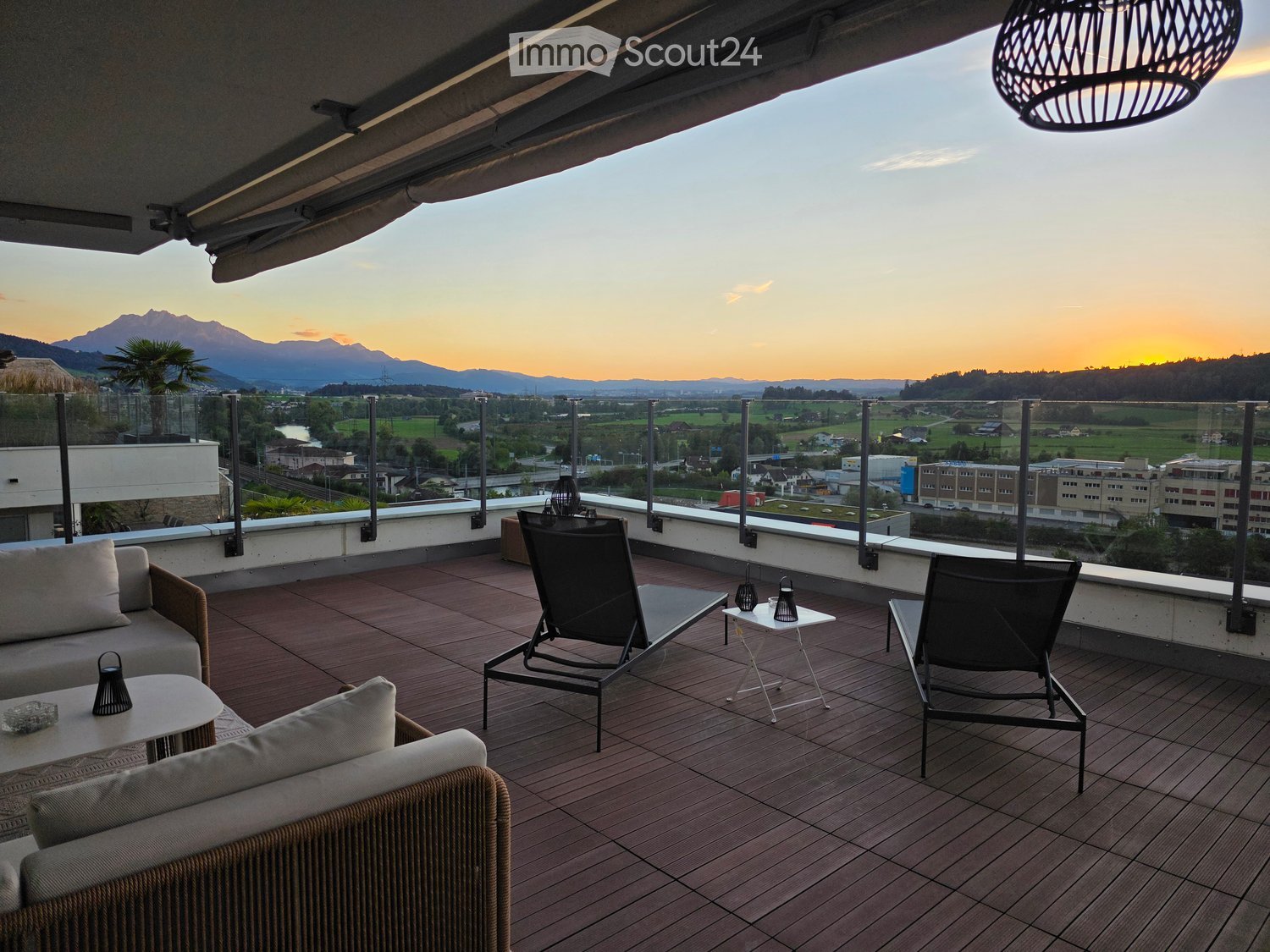 Balcony with wooden decking, sofa, chairs, small tables, and a scenic mountain view at sunset.