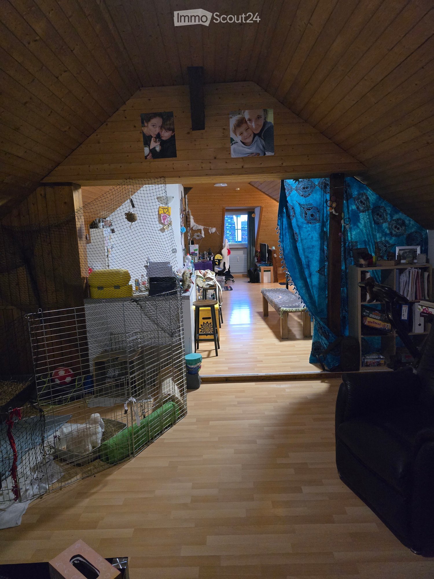 A spacious, wooden-floored room with a high, slanted ceiling. Features include a black sofa on the right, a small stool near the center, a pet cage with a ball on the left, and a curtained area with shelves and a table. The room is well-lit with natural l
