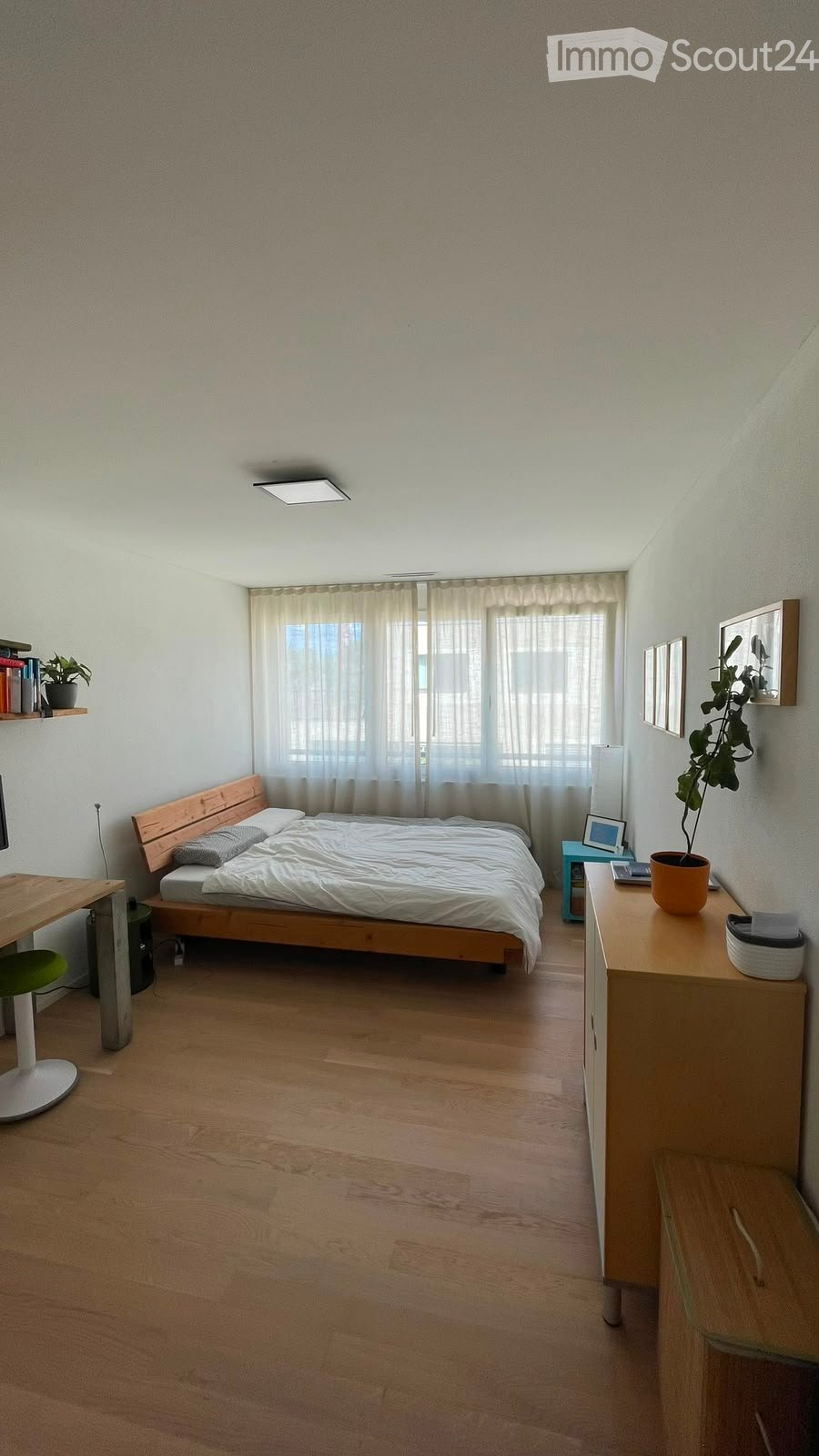Cozy bedroom with wooden flooring, a bed with white bedding, a nightstand with a lamp, a desk with a chair, and a shelf with books and a plant.