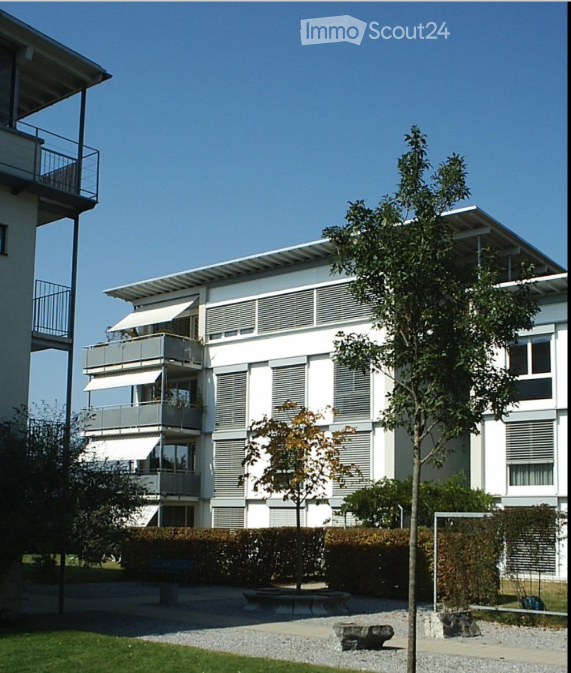 Modern multi-story residential building with white facade, large windows, and balconies. The building features a flat roof and is surrounded by a landscaped area with trees, shrubs, and a stone bench.