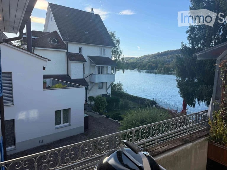 Balcony overlooking a serene lake with surrounding greenery and a multi-story house in the background.