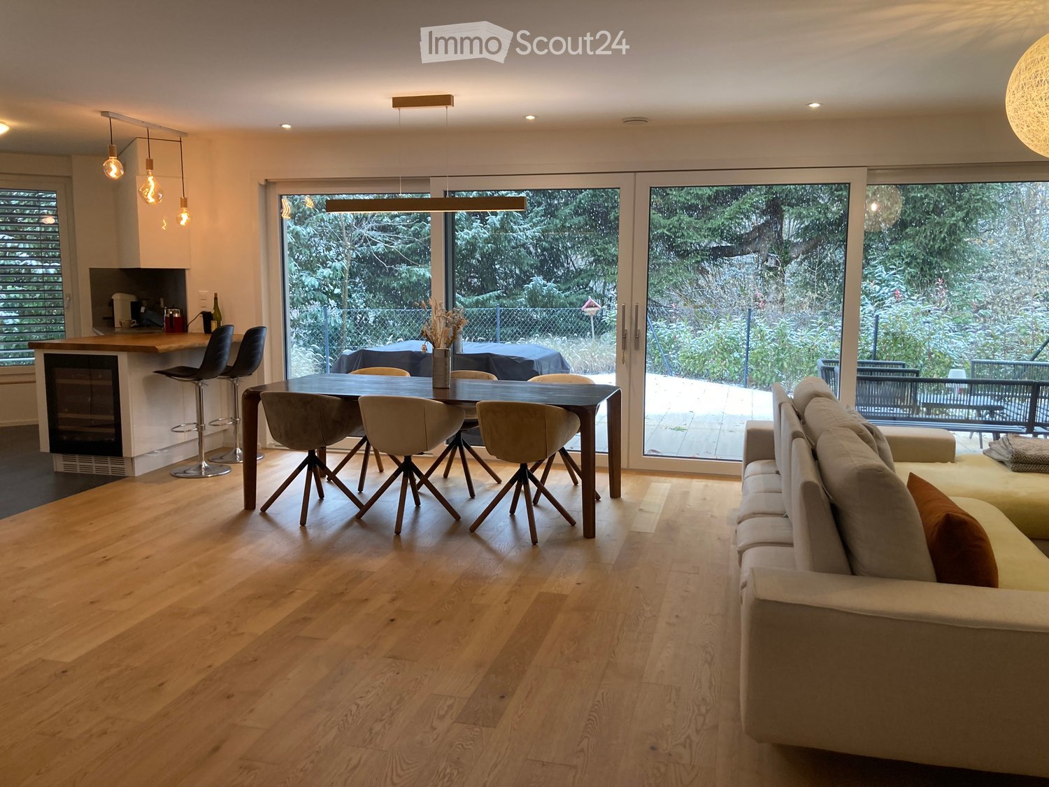 Open-plan living and dining area with light wooden flooring, a large sofa, a dining table with chairs, and a kitchen island with bar stools. Large windows offer a view of the garden.