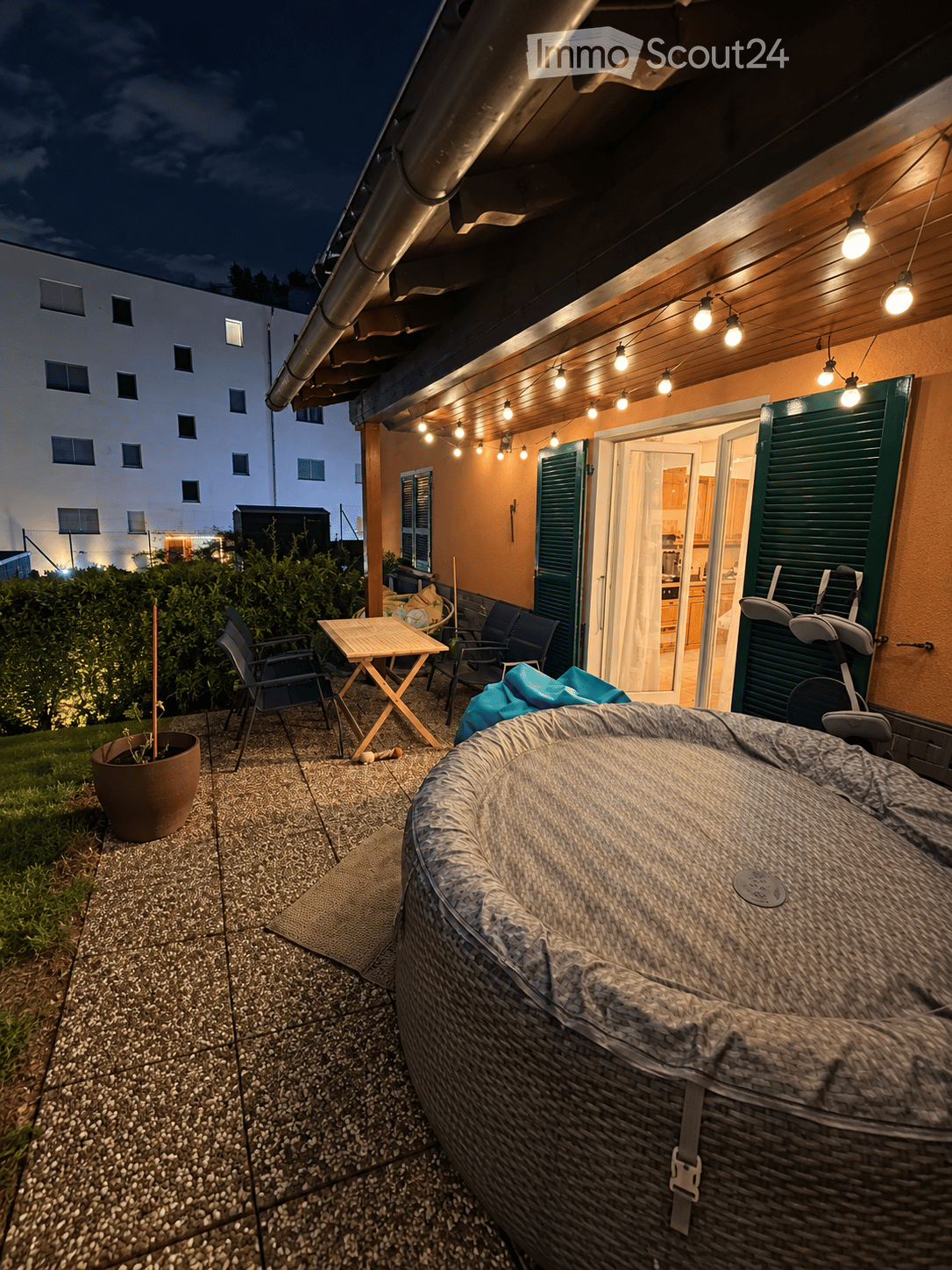 Outdoor patio area with a hot tub, small table, chairs, and a potted plant, illuminated by string lights.