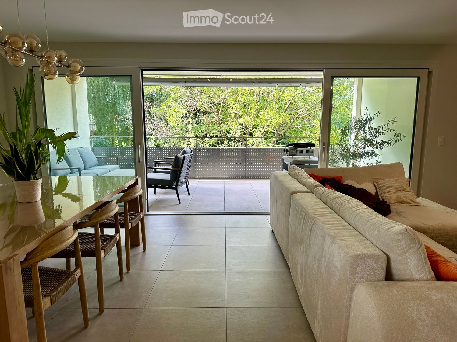 Modern living room with a large sofa, glass dining table, wooden chairs, and a potted plant near the sliding glass doors leading to a balcony.