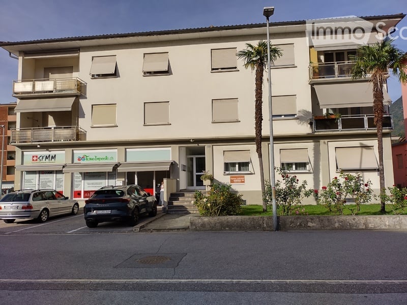 Three-story building with a modern facade, multiple windows with shutters, and a small entrance area. There are two cars parked in front, and the building has a small garden area with palm trees and shrubs.