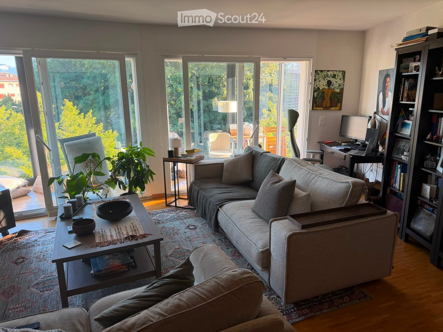 Modern living room with large windows offering a scenic view, featuring a grey sectional sofa, coffee table, side table, and a bookshelf with books and decorative items.