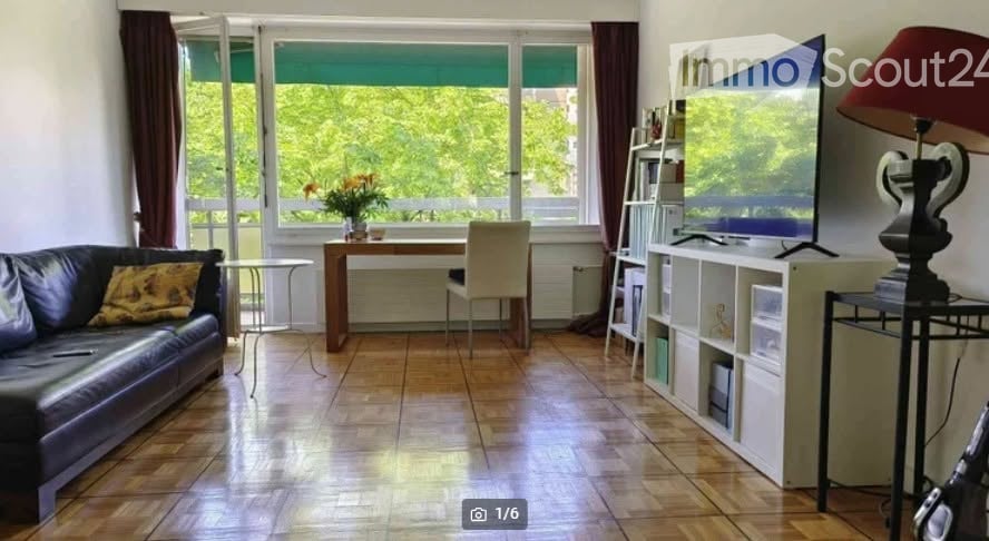 Bright living room with large windows offering a view of greenery, featuring a black leather sofa, wooden coffee table, white chair, bookshelf, and a television on a stand.
