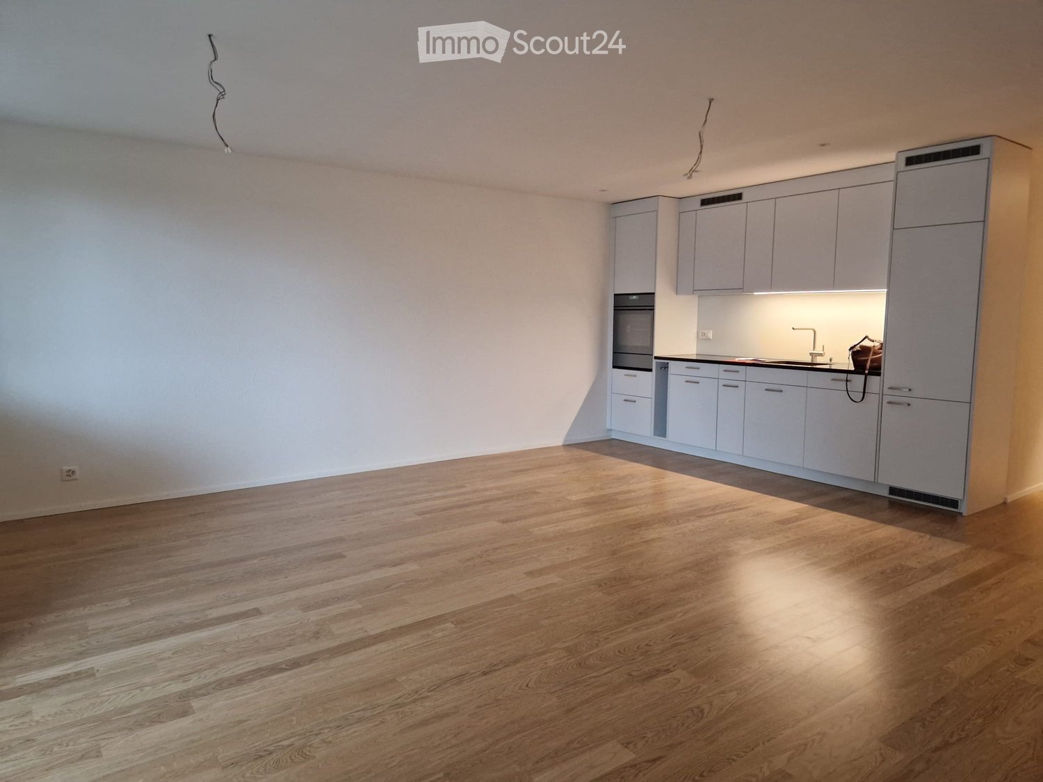Empty room with light wooden flooring and white walls, featuring a modern kitchen area with cabinets and appliances.