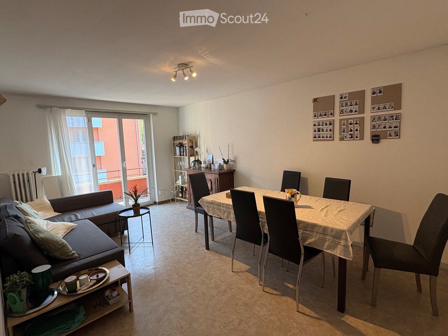 A modern living and dining area with a sofa, coffee table, dining table with chairs, and a bookshelf. The room features large windows with a sliding glass door leading to a balcony.