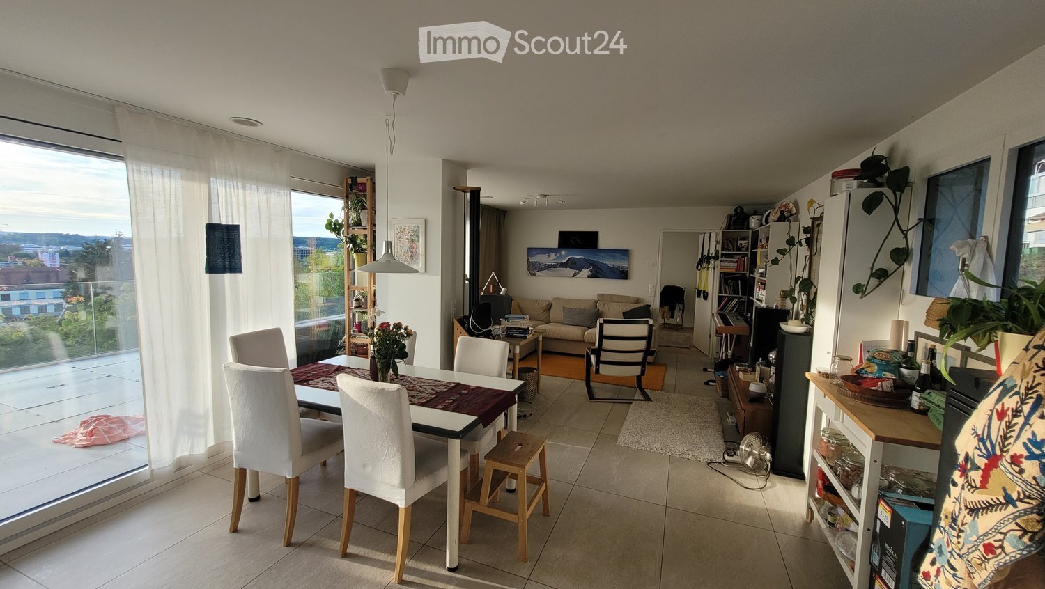 Bright, open-plan living and dining area with large windows and a view, featuring a dining table with chairs, a sofa, a small table, and various decorative items.