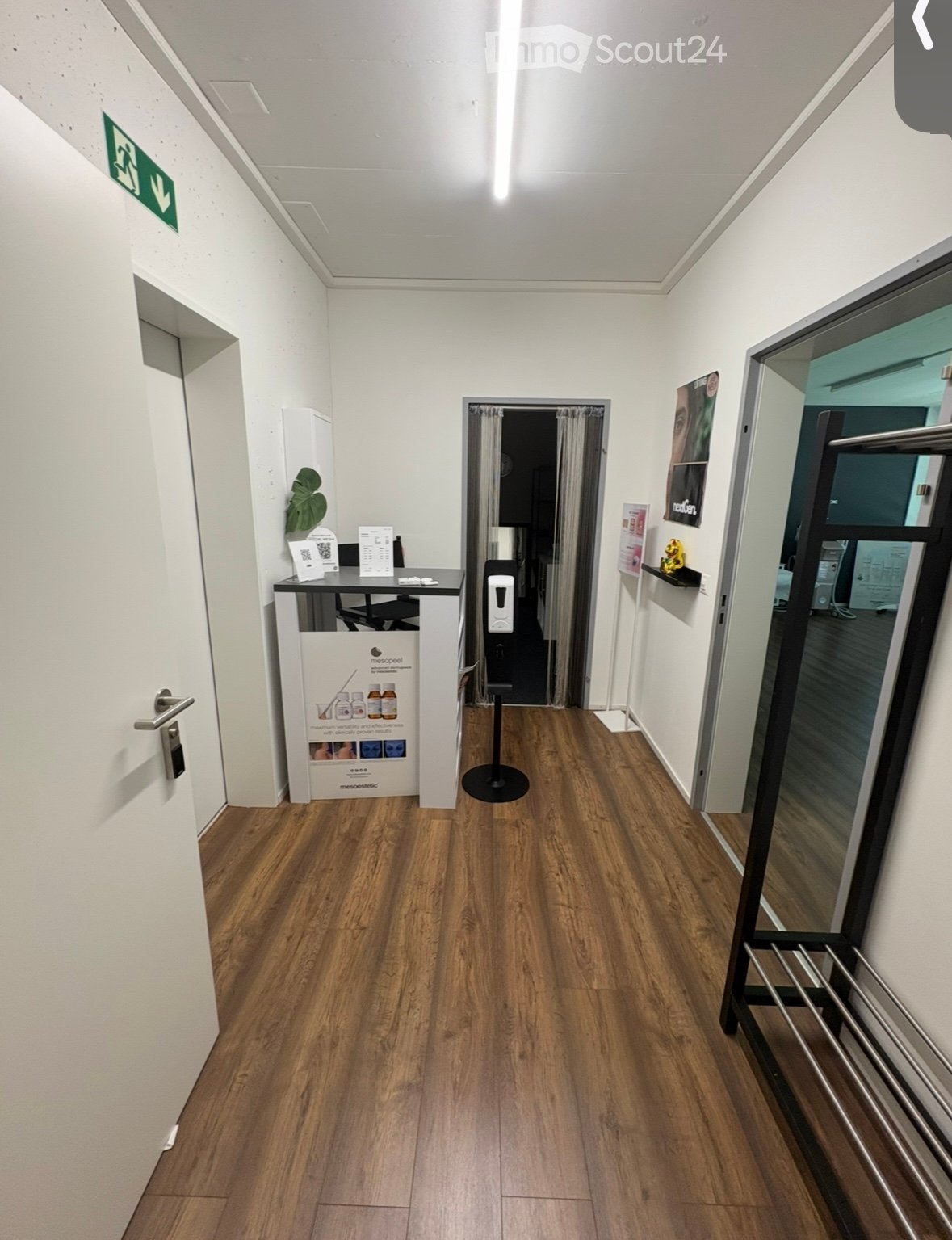 A clean, well-lit hallway with wooden flooring, white walls, and a reception area visible on the left side. There is a door with a silver handle, a small table with brochures, and a poster on the wall. A glass door with a metal frame is visible on the rig