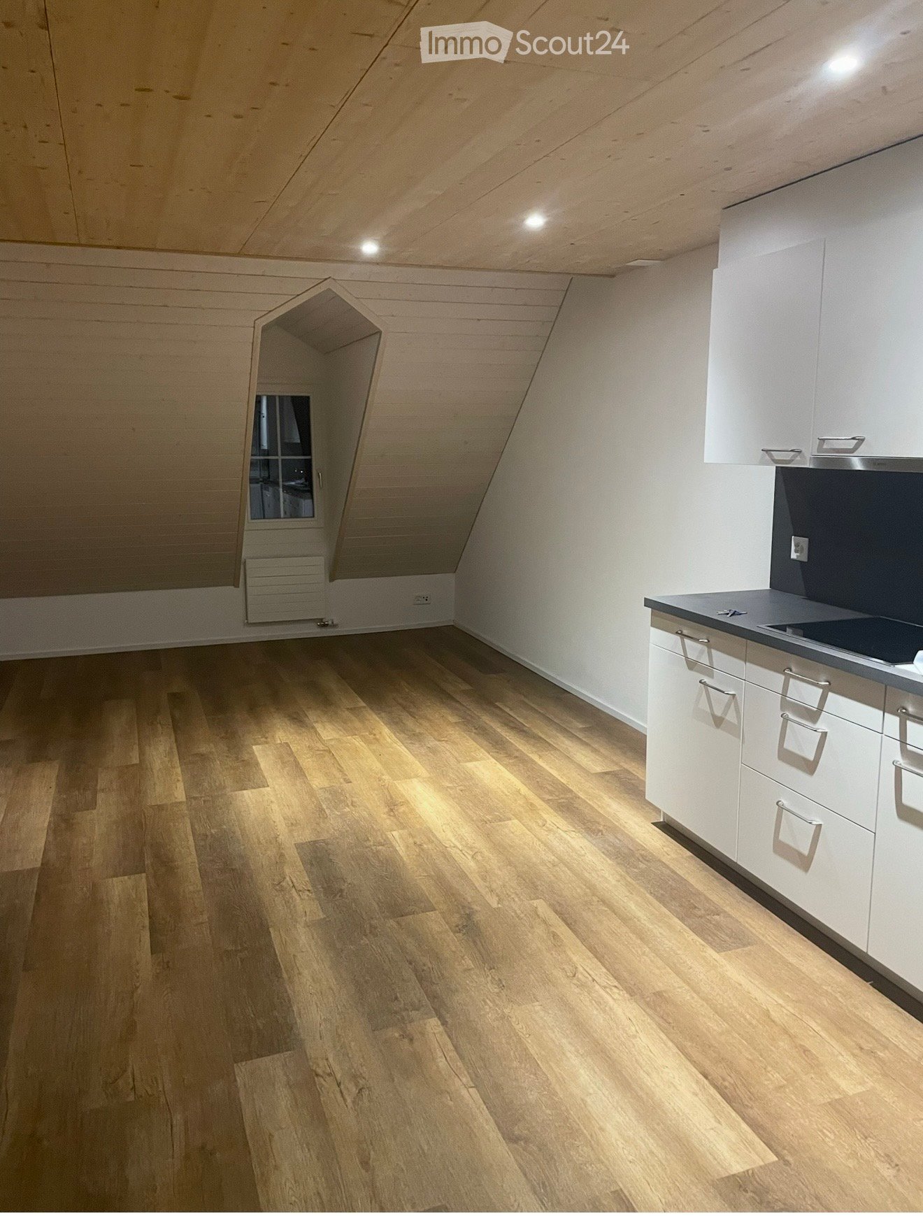 An unfurnished attic room with wooden flooring, white walls, and a small window. The room features built-in white cabinets and a countertop on the right side.