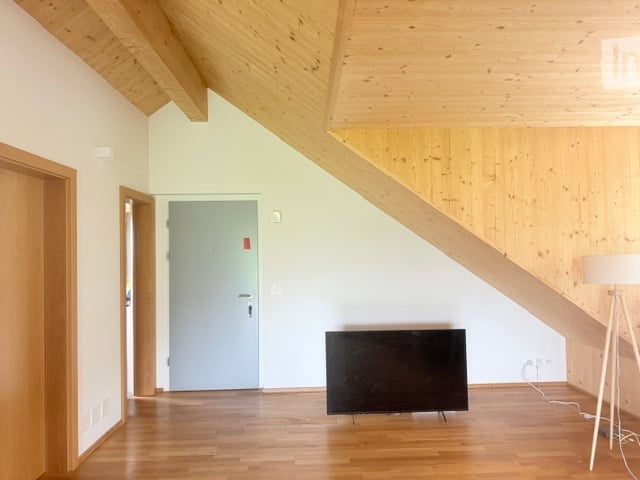 Bright, wooden-floored room with exposed wooden beams on the ceiling, featuring a flat-screen TV on a stand and a modern floor lamp.