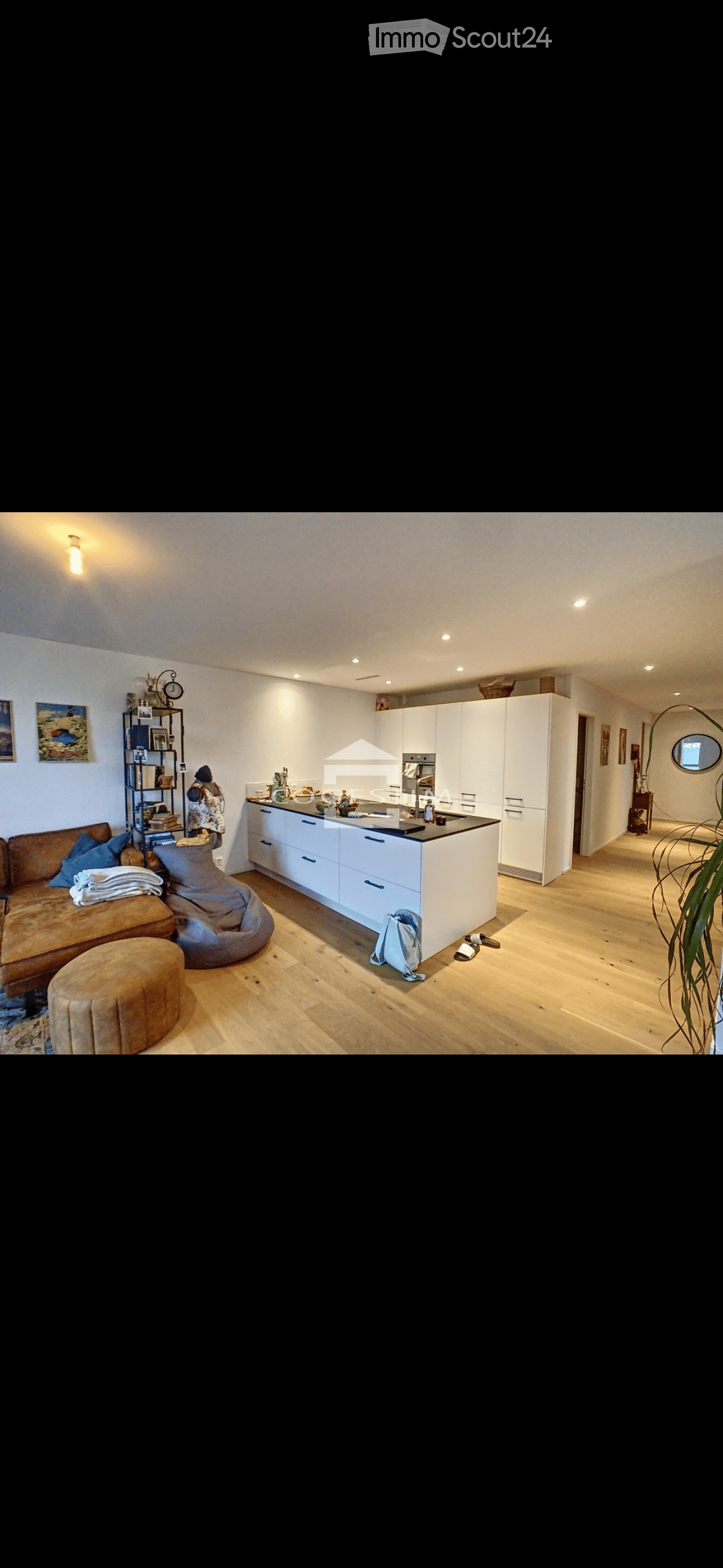 Open-plan living and kitchen area with a sofa, a round stool, a kitchen island, white cabinets, a shelf with books and decor, and a large plant near the entrance.